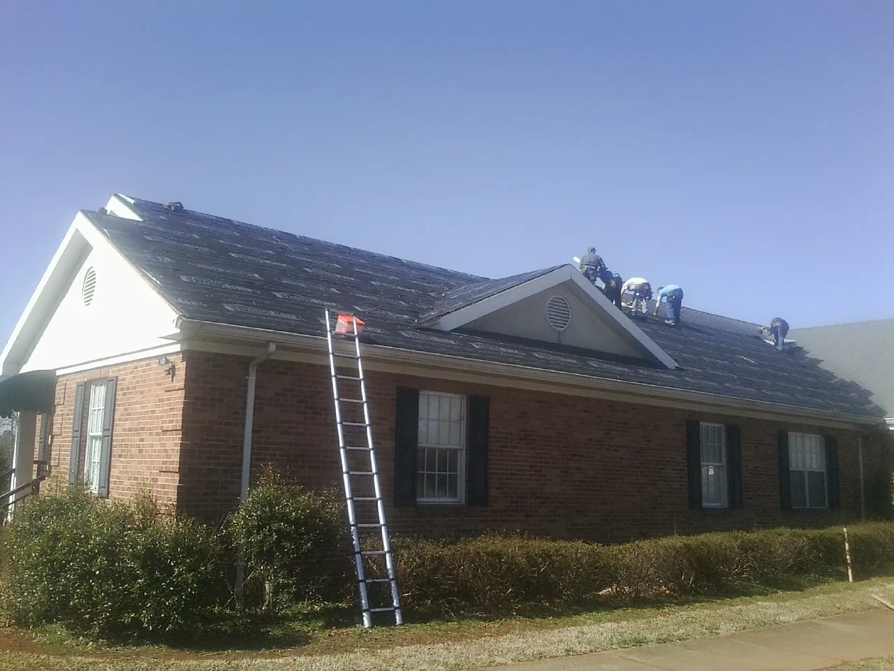 Crew tearing shingles off a brick ranch roof with a ladder set — Emergency Roof Repair work by our crew in East Leroy
