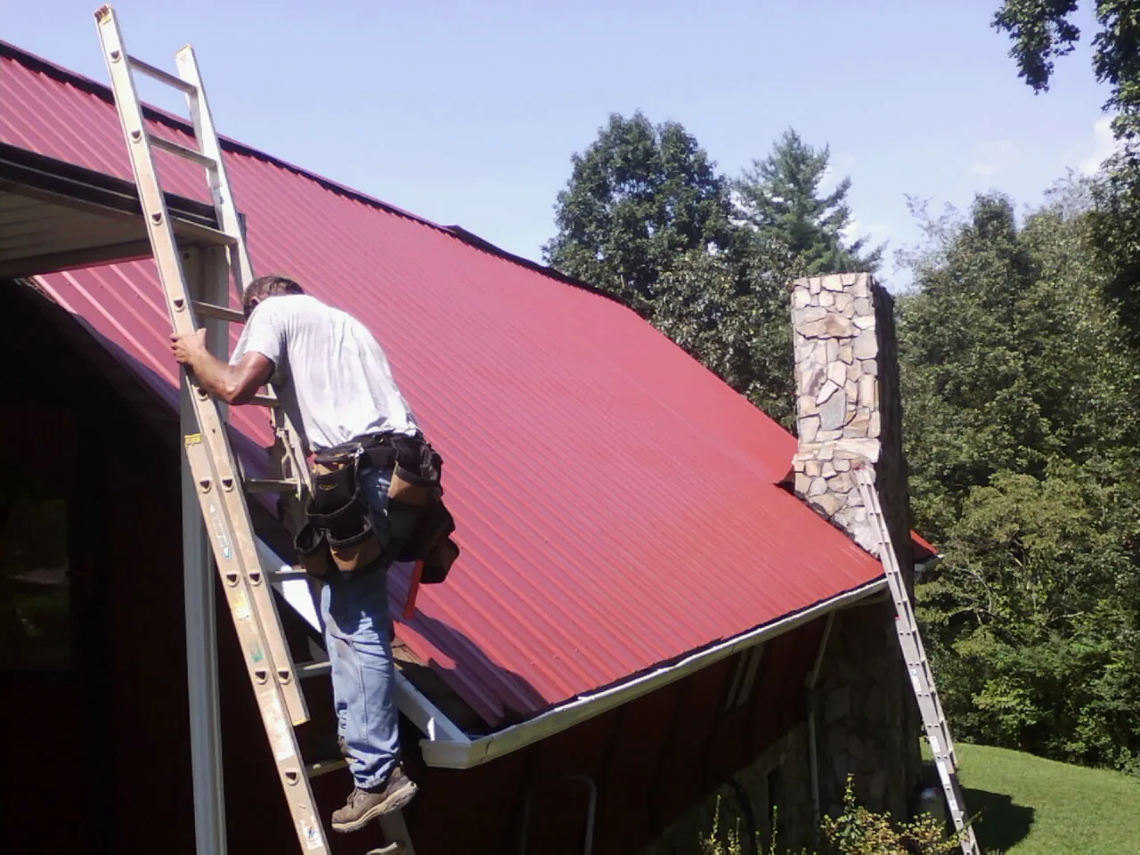 Roofer on a ladder beside a red metal roof and stone chimney — Soffit & Fascia Repair by Roofing Brothers in East Leroy